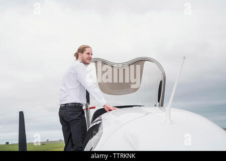 Side view of confident male pilot looking away while standing by airplane against cloudy sky Stock Photo