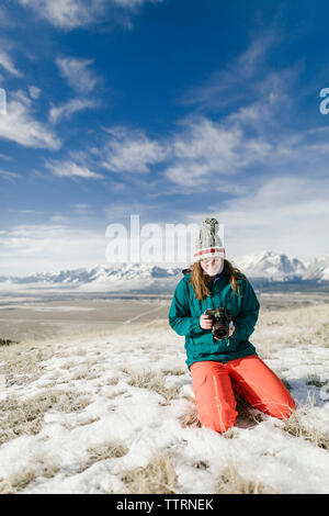 Female hiker looking at camera while kneeling on snowy field against mountains Stock Photo