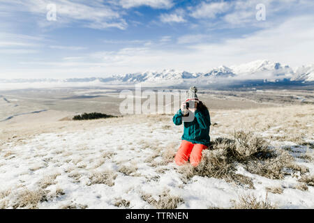 Female hiker photographing with camera while kneeling on snowy field against mountains Stock Photo