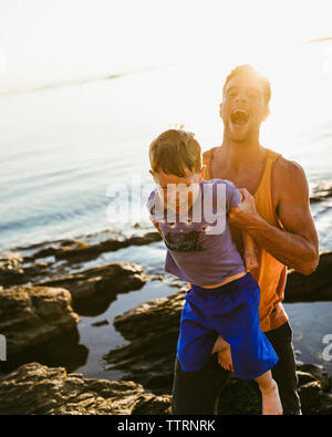 Happy father carrying son while standing on rocks at beach against sky during sunset Stock Photo