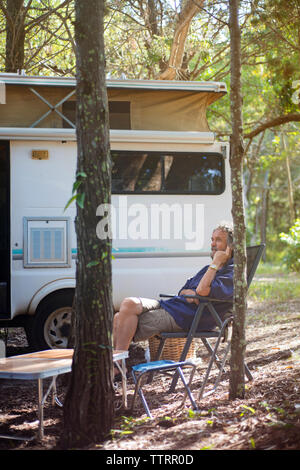 Thoughtful senior man sitting on folding chair against travel trailer in forest Stock Photo