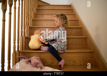 Side view of girl playing with doll while sitting on wooden steps at home Stock Photo