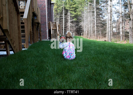Rear view of girl holding American football while sitting on grassy field in yard Stock Photo
