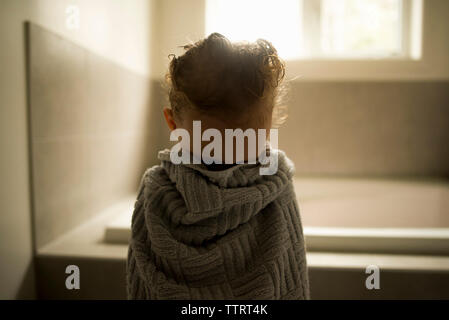 Girl wrapped in towel hiding face while standing at bathroom Stock Photo