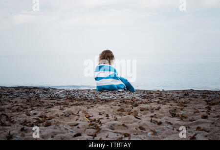Rear view of girl wrapped in towel sitting at lakeshore against sky Stock Photo