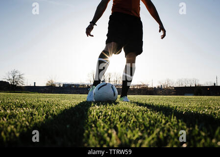 Low section of man playing soccer on grassy field against clear sky during sunset Stock Photo