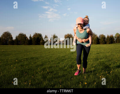 Happy mother piggybacking daughter while exercising on grassy field against sky at park Stock Photo