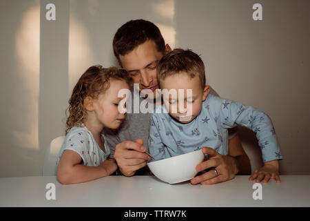 Father feeding children while sitting by table against wall at home Stock Photo