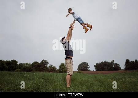 Happy father throwing son in air while standing on grassy field against sky at park Stock Photo