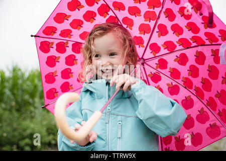 Happy cute girl carrying pink umbrella while standing outdoors during rainy season Stock Photo