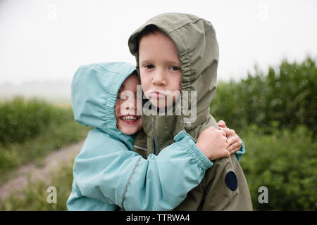 Portrait of sad brother being embraced by happy sister while standing against plants during rainy season Stock Photo