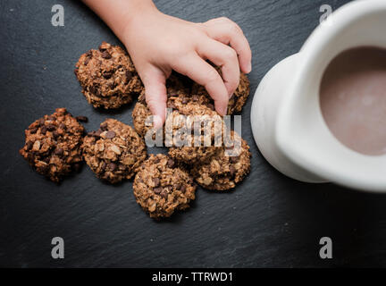 Cropped hand of girl picking chocolate chip cookies from slate by coffee in jug Stock Photo