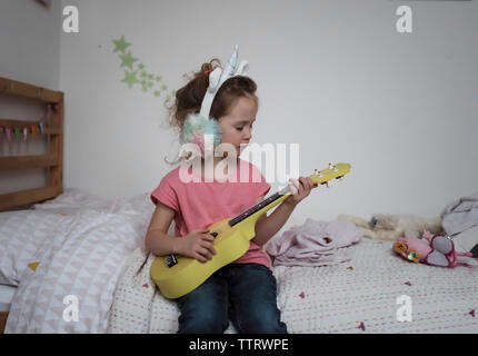 young girl playing ukulele wearing face dress as unicorn in bedroom Stock Photo