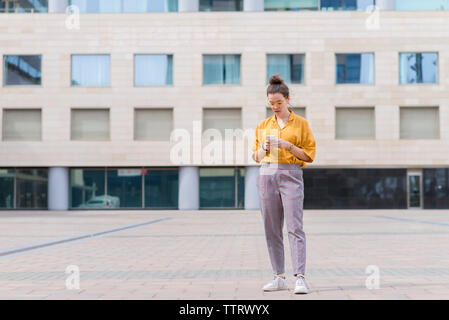 Confident businesswoman using smart phone while standing on sidewalk against building in city Stock Photo