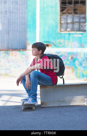 Side view of thoughtful student with skateboard carrying backpack while sitting in city Stock Photo