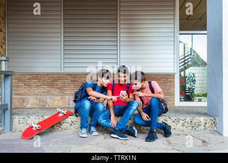 Friends using smart phone while sitting against school building Stock Photo