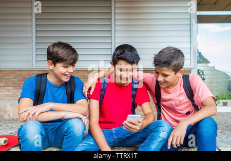Happy friends using smart phone while sitting against school building Stock Photo