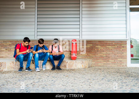 Friends using smart phones while sitting against school building Stock Photo
