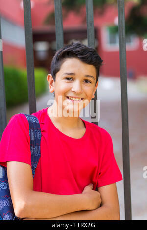 Portrait of smiling schoolboy with backpack standing by gate against school building Stock Photo
