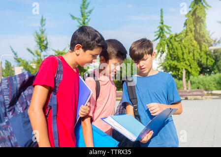 Schoolboys discussing while standing on footpath against sky during sunny day Stock Photo