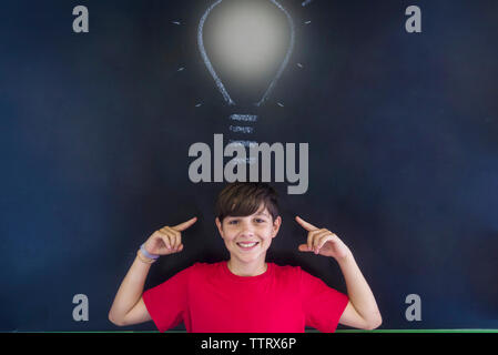 Portrait of smiling schoolboy with light bulb drawing on blackboard standing in classroom Stock Photo