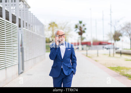 Confident senior man in suit talking through smart phone while walking on footpath against sky Stock Photo