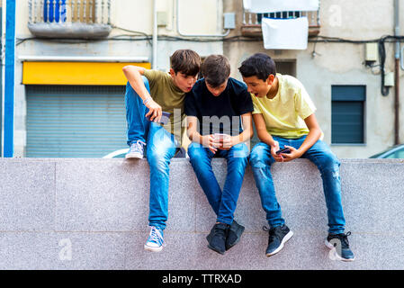 Surprised boys looking at friend using smart phone while sitting on retaining wall against building in city Stock Photo