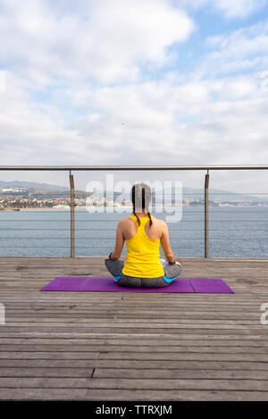 Rear view of woman in sportswear sitting on mat, stretching on pier Stock Photo