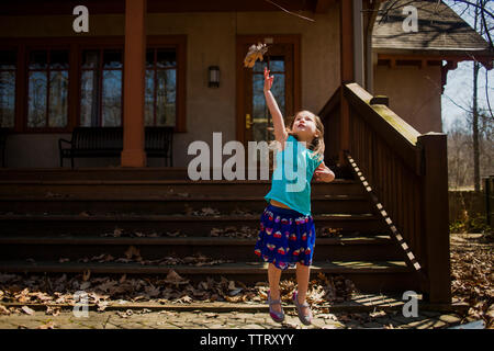 Playful girl jumping while throwing dry leaf in air against house during autumn Stock Photo