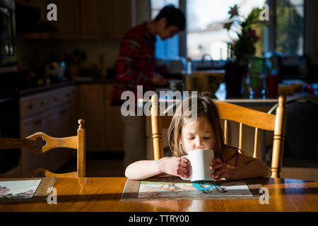Daughter drinking tea on table while father working in background at home Stock Photo