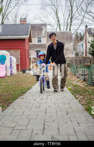 Happy father assisting son in riding bicycle on footpath at backyard Stock Photo