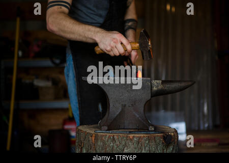 Midsection of blacksmith forging knife on anvil in workshop Stock Photo