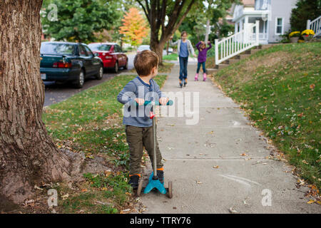 Brother riding push scooter while sisters skating on footpath in background Stock Photo