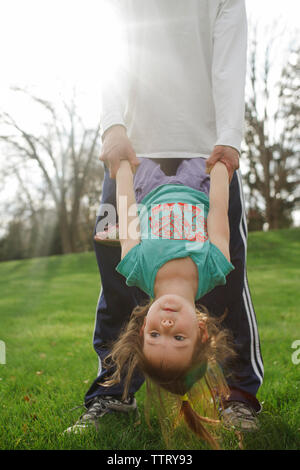 Low section of father holding daughter upside down while standing on grassy field against sky at park Stock Photo