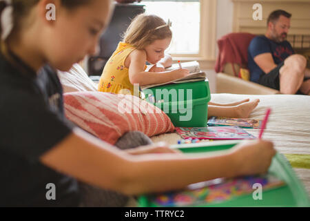 Sisters drawing while sitting on bed against father at home Stock Photo