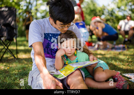 Father with daughter reading magazine while sitting on grassy field in park Stock Photo
