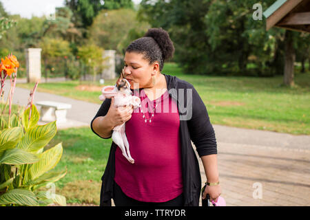 Woman kissing Chihuahua while standing on footpath against trees in park Stock Photo