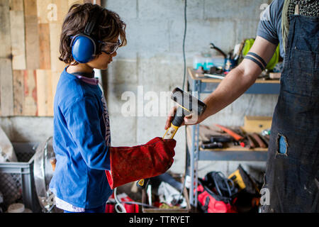 Midsection of blacksmith giving hammer to boy while standing against wall in workshop Stock Photo