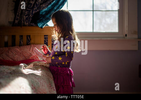 A small girl stands in patch of light in bedroom writing in journal Stock Photo
