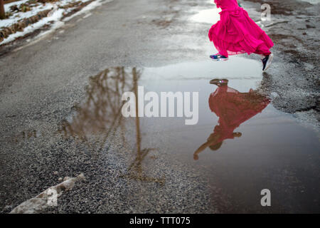 Reflection of little girl leaping joyfully in long dress across puddle Stock Photo