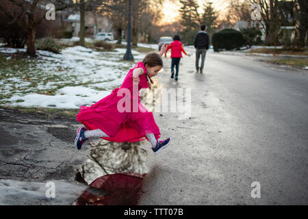 A little girl in long dress leaps across puddle on walk with family Stock Photo