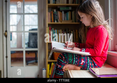 side view of a little girl drawing pictures in her pajamas by window Stock Photo