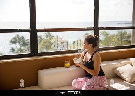 Woman looking through window while eating breakfast on sofa at home Stock Photo