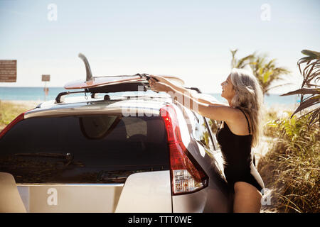 Side view of mature woman in wetsuit tying surfboard on car roof at Delray beach Stock Photo