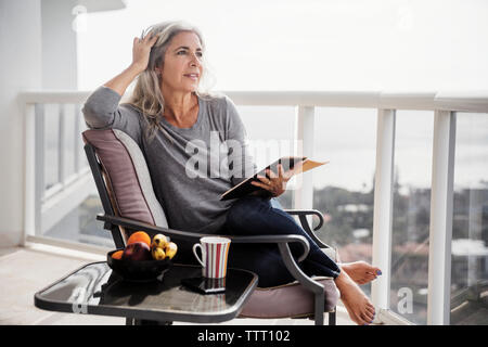 Thoughtful woman holding book while sitting on chair on balcony Stock Photo