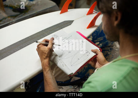 worker writing in book while analyzing surfboard in workshop Stock Photo
