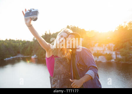 Friends taking selfie through camera by lake during sunset Stock Photo