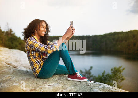 Happy woman taking selfie through smart phone on rock by lake Stock Photo