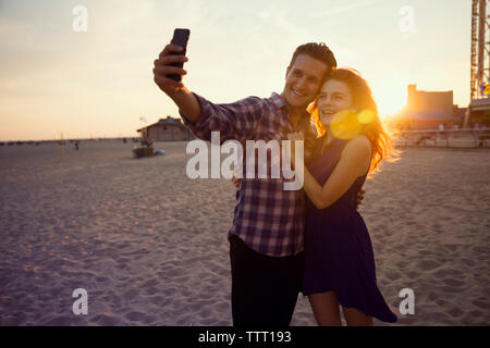 Young couple taking selfie while standing on sand during sunset Stock Photo
