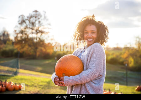 Portrait of smiling woman carrying pumpkin while standing in farm against sky Stock Photo